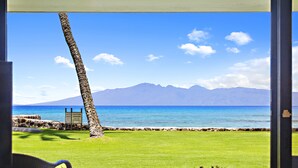 A view from a window overlooking a beach with a grassy area, a palm tree, and mountains in the background. - Premier Spacious Condo with Stunning Ocean Views (Lahaina)