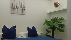 Quarto com cama, almofadas, pintura na parede, uma planta pequena e uma prateleira com uma flor.