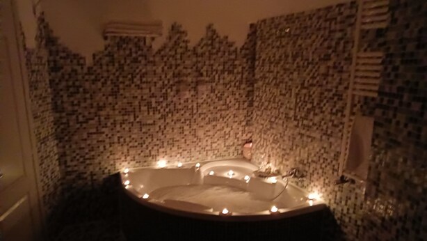 Um quarto com iluminação fraca, apresentando uma banheira grande e redonda cheia de água e velas. As paredes são revestidas com um padrão de mosaico.