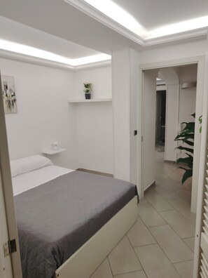 A bedroom with a bed, a bedside table, a wall-mounted shelf with a plant, and a door leading to another room.