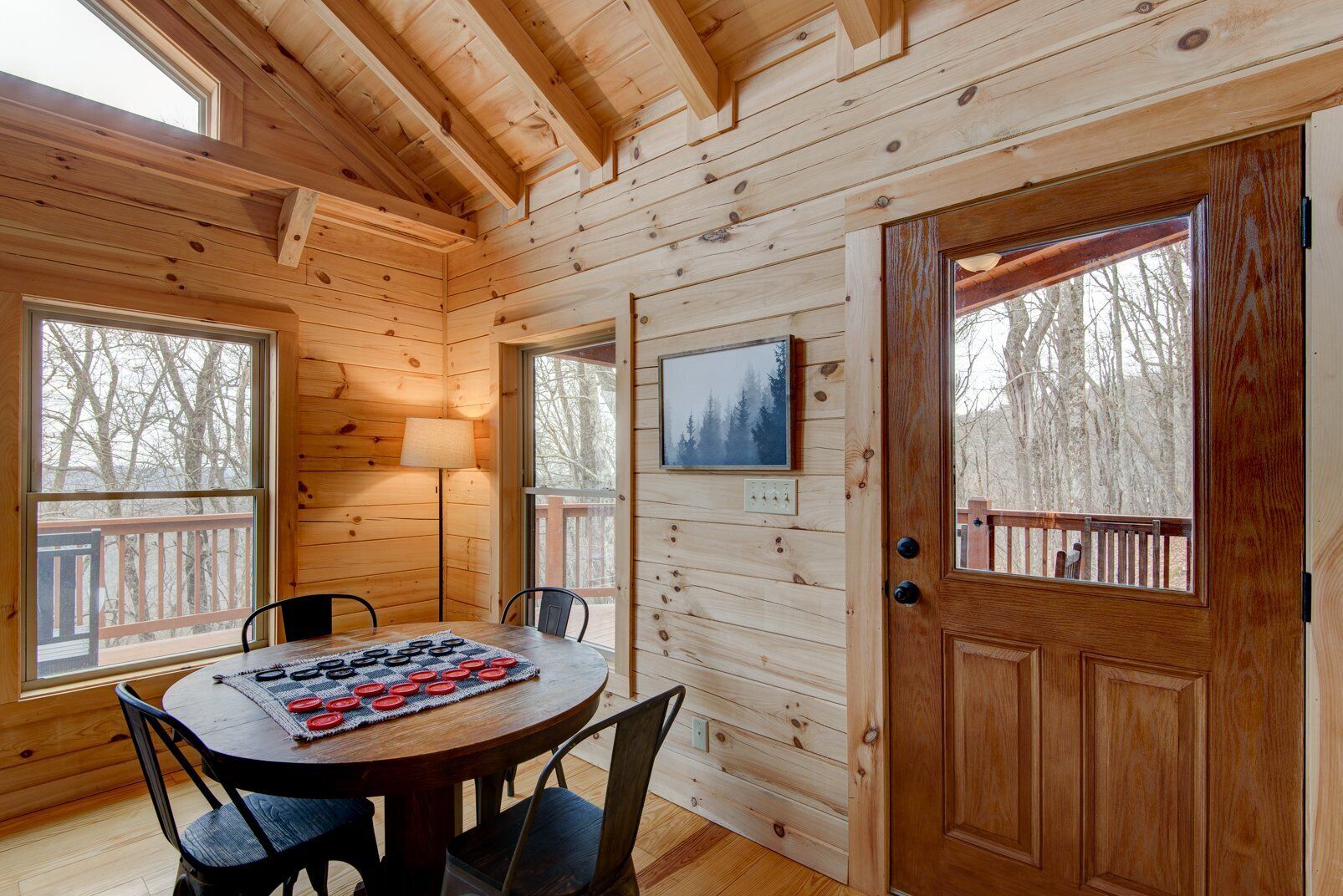 A wooden cabin interior with a dining table, chairs, a lamp, and a door leading outside.