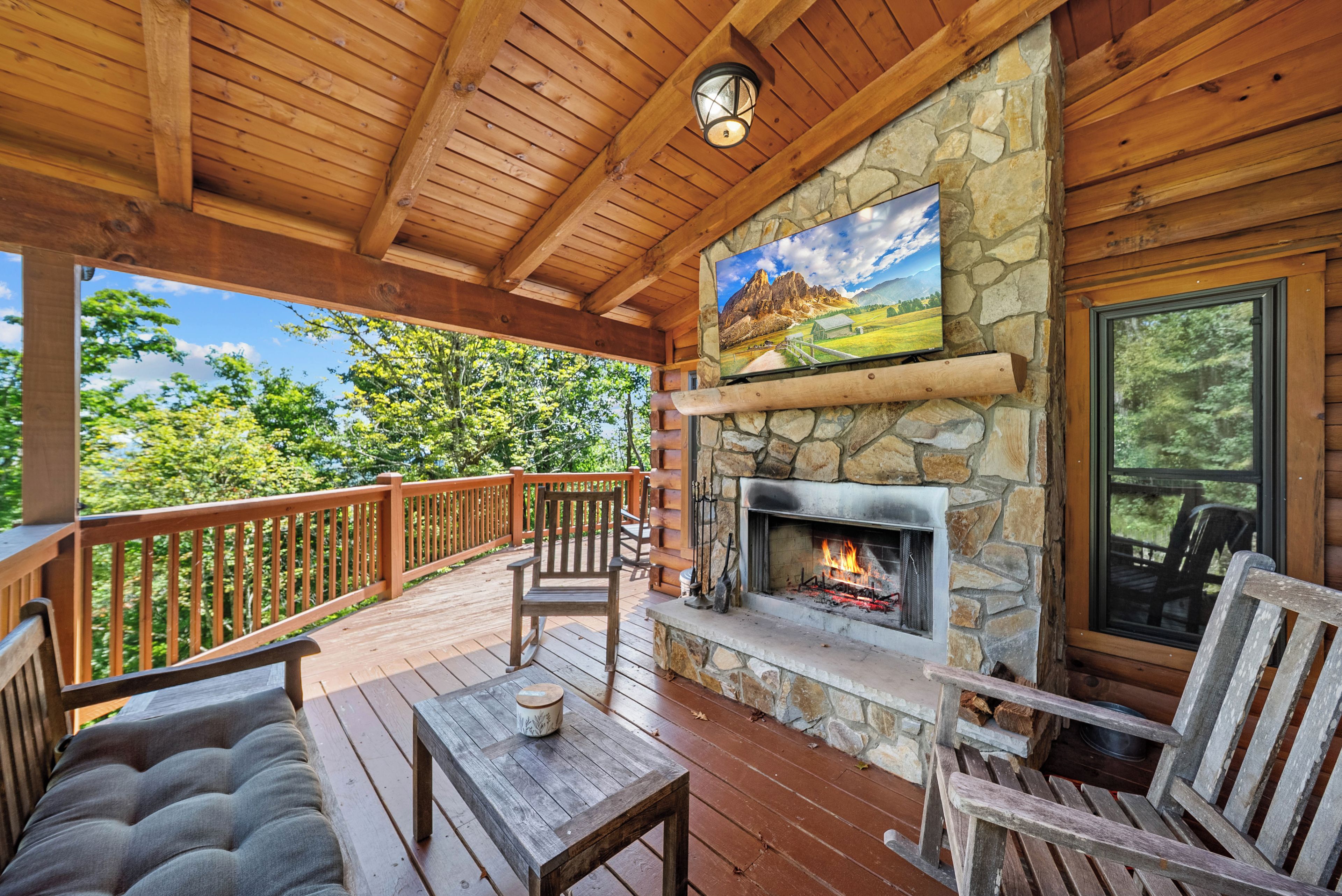 A wooden deck with a stone fireplace, a mounted television, and outdoor seating.