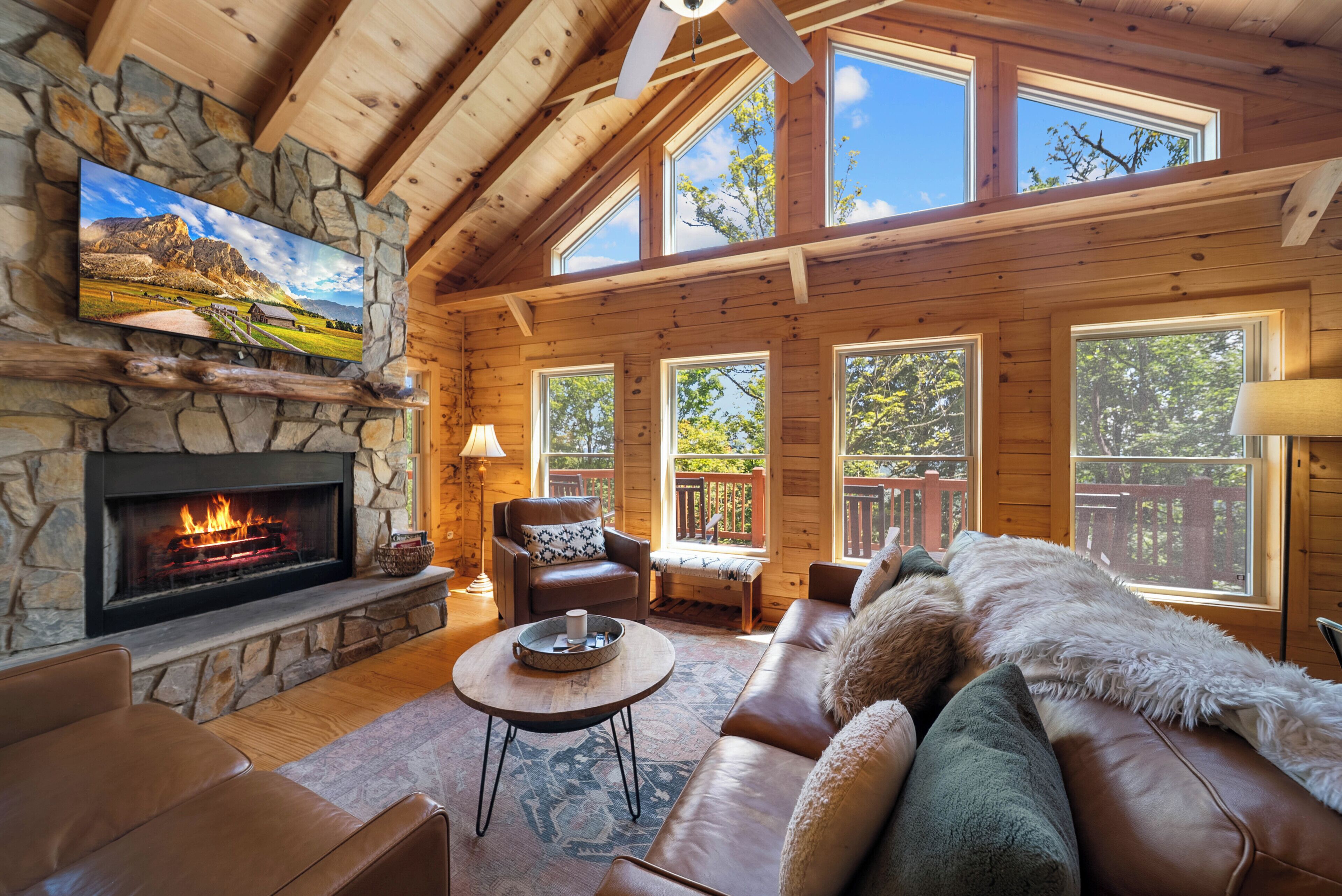 A cozy cabin interior with a stone fireplace, leather furniture, and large windows offering views of the outdoors.