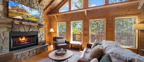 A cozy cabin interior with a stone fireplace, leather furniture, and large windows offering views of the outdoors.