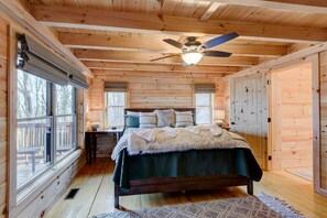 Un dormitorio de cabaña de madera con una cama, dos ventanas, un ventilador de techo y una terraza de madera afuera.