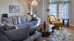 Un salon avec un canapé bleu, un fauteuil jaune, une table basse en bois et une grande fenêtre avec des stores.
