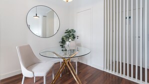 A modern dining area with a round glass table, two chairs, a mirror, and a hanging light fixture. - GuestReady - Almada 516 (Porto)