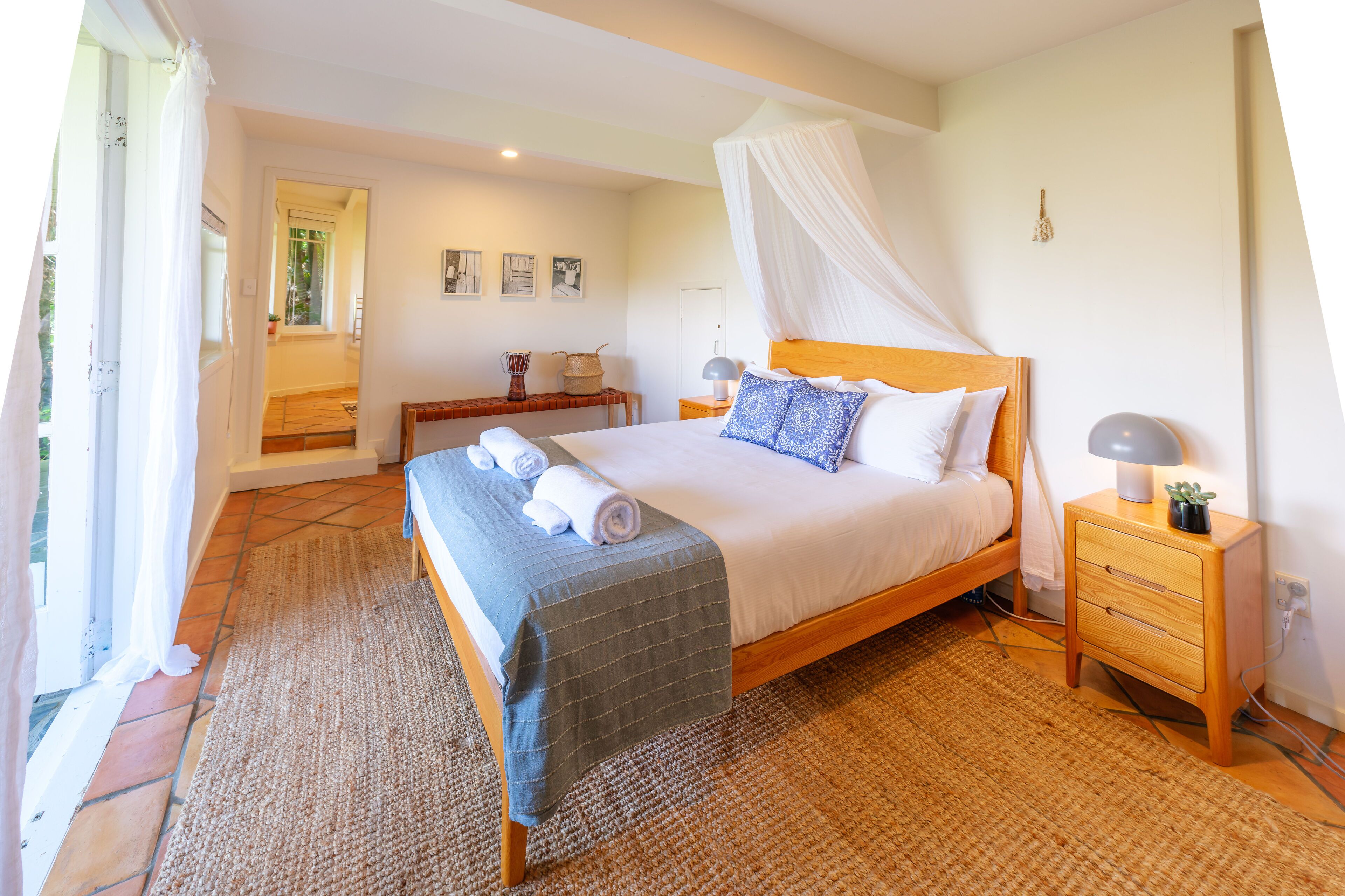 A bedroom with a canopy bed, a bedside table with a lamp, and a view of the outside.