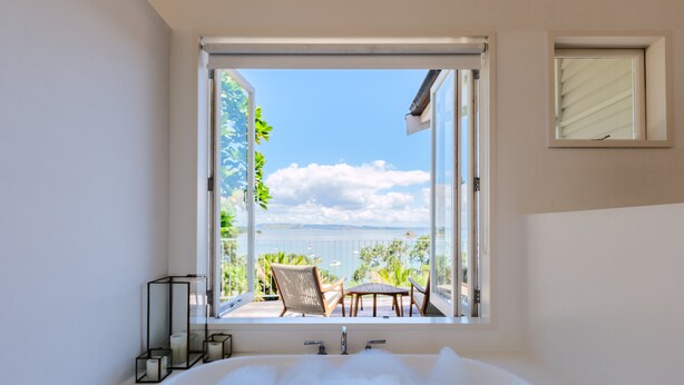 A bathroom with a large bathtub, a window with a view of a balcony, and a small window on the wall. - Koi Waiheke with private beach access (Waiheke Island)
