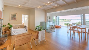 A spacious living area with a sofa, a dining table with chairs, and a view through large windows. - Koi Waiheke with private beach access (Waiheke Island)