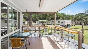A covered outdoor seating area with tables and chairs, a view of a campsite with RVs, and a building with large windows. - Shoalhaven Heads Village (Shoalhaven Heads)