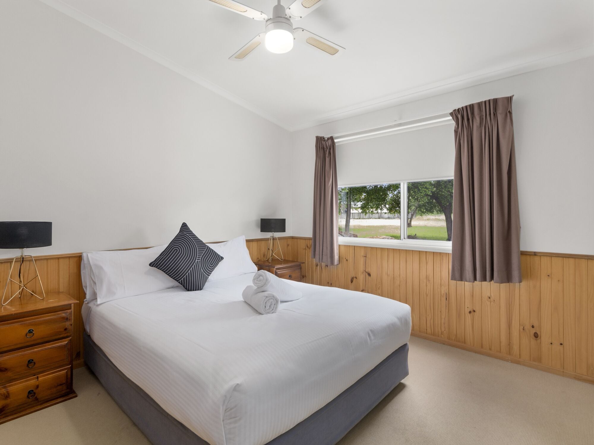 Une chambre à coucher avec un lit, des murs recouverts de panneaux de bois, une fenêtre avec des rideaux, un ventilateur de plafond et des lampes de chevet.