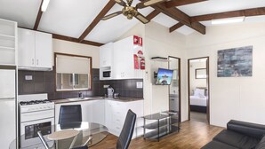 A modern kitchen with white cabinets, a gas stove, and a microwave. There is a dining area with a glass table and chairs. The room has wooden flooring and a ceiling fan. - Shoalhaven Heads Village (Shoalhaven Heads)