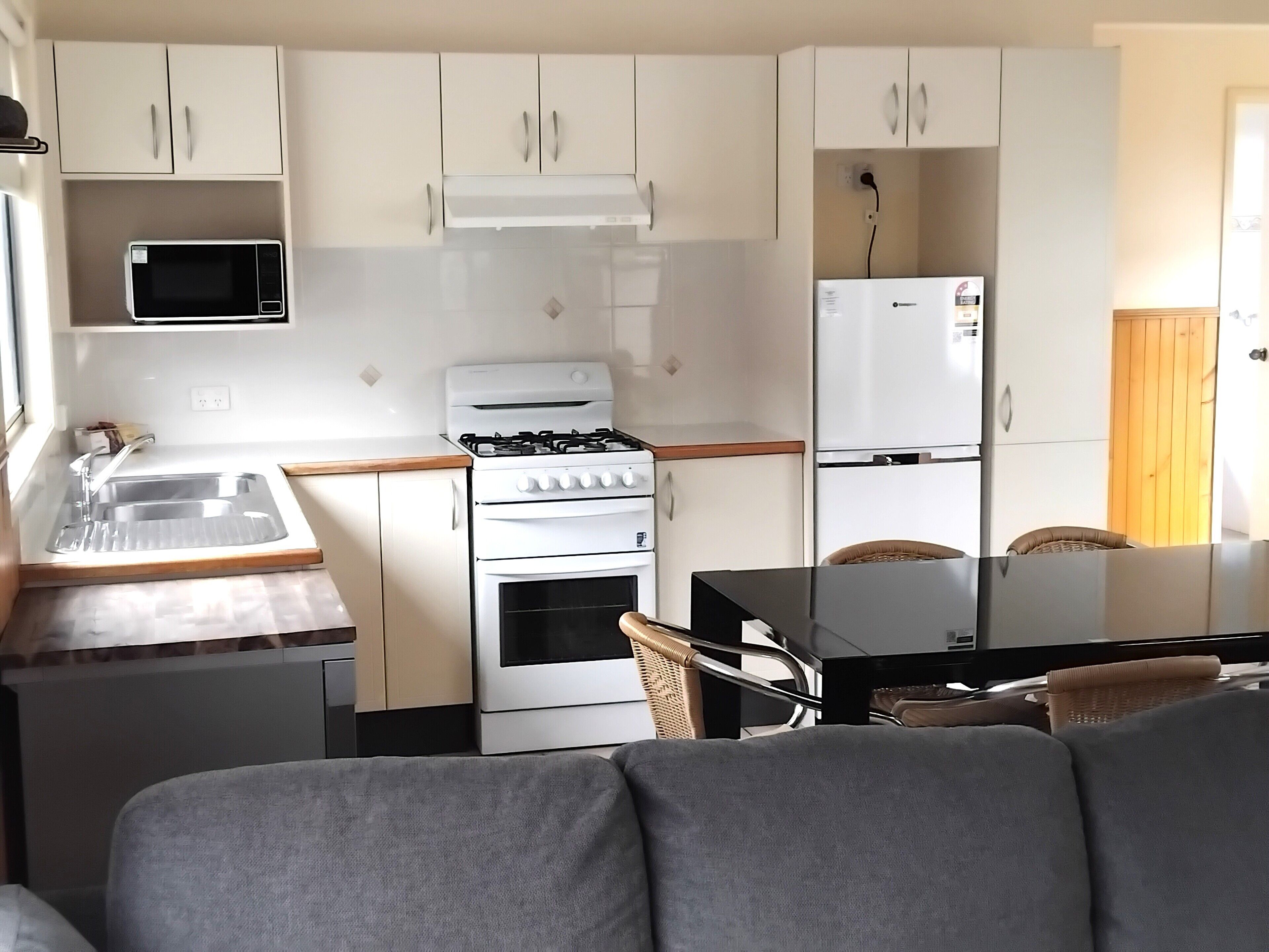 A compact kitchen with white cabinets, a microwave, a stove, and a refrigerator. There is a dining table with chairs and a sofa in the foreground.