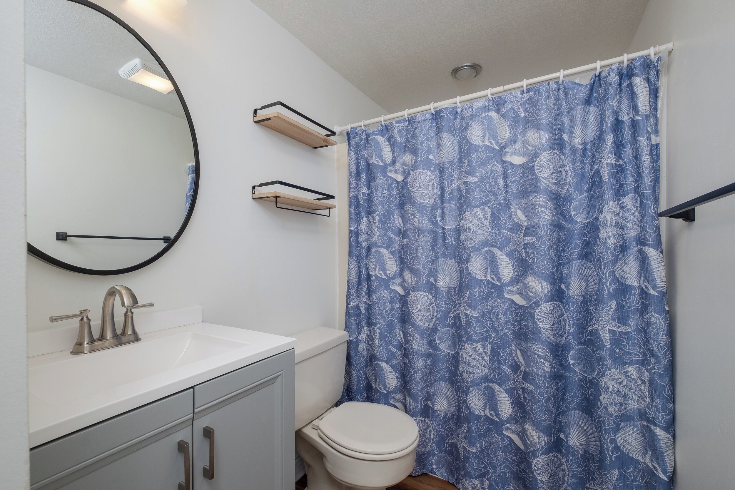 A bathroom with a white sink, a round mirror, a toilet, and a blue shower curtain with a shell pattern.