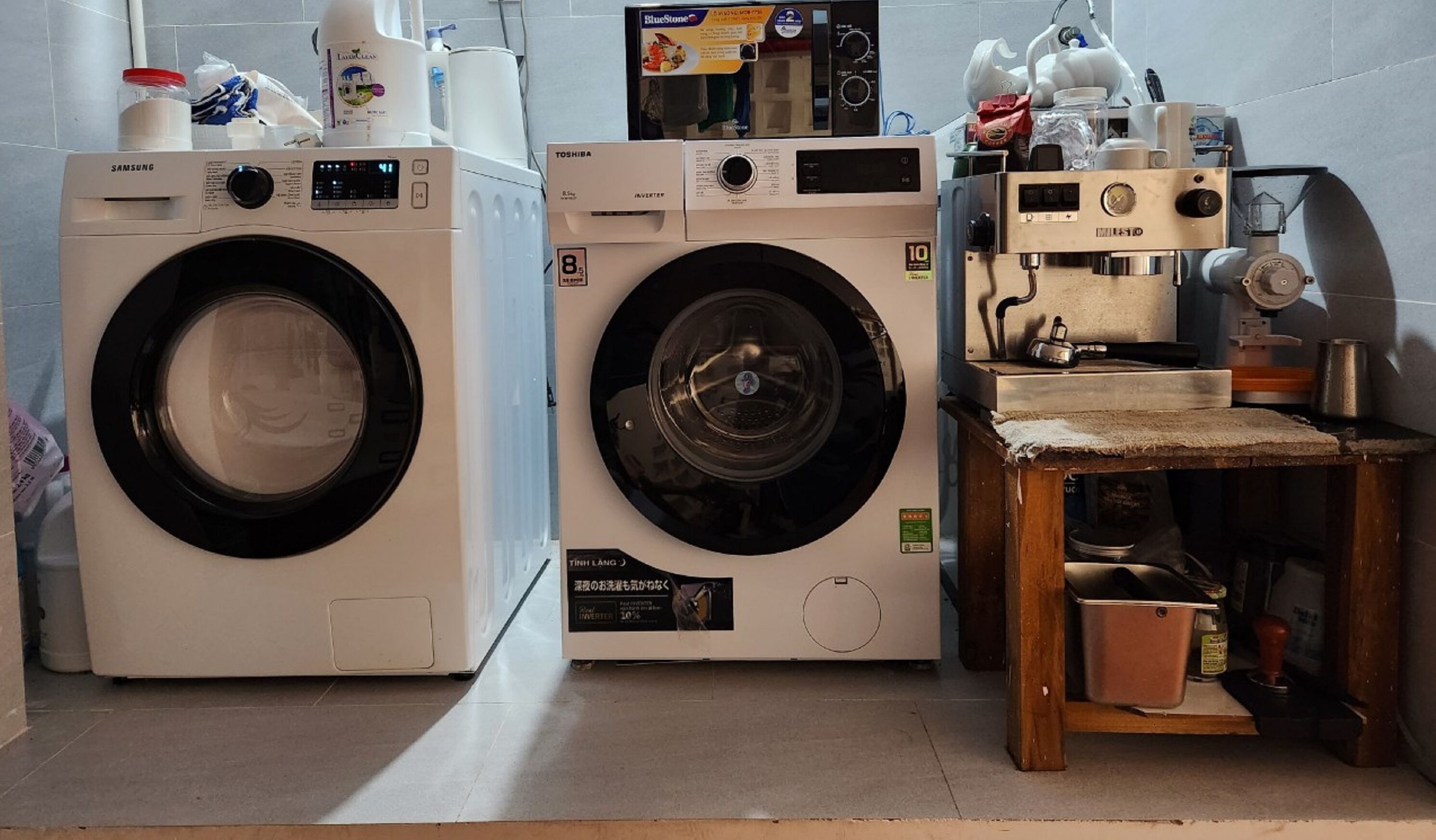 Two front-loading washing machines in a laundry area with a small table and various items on top.