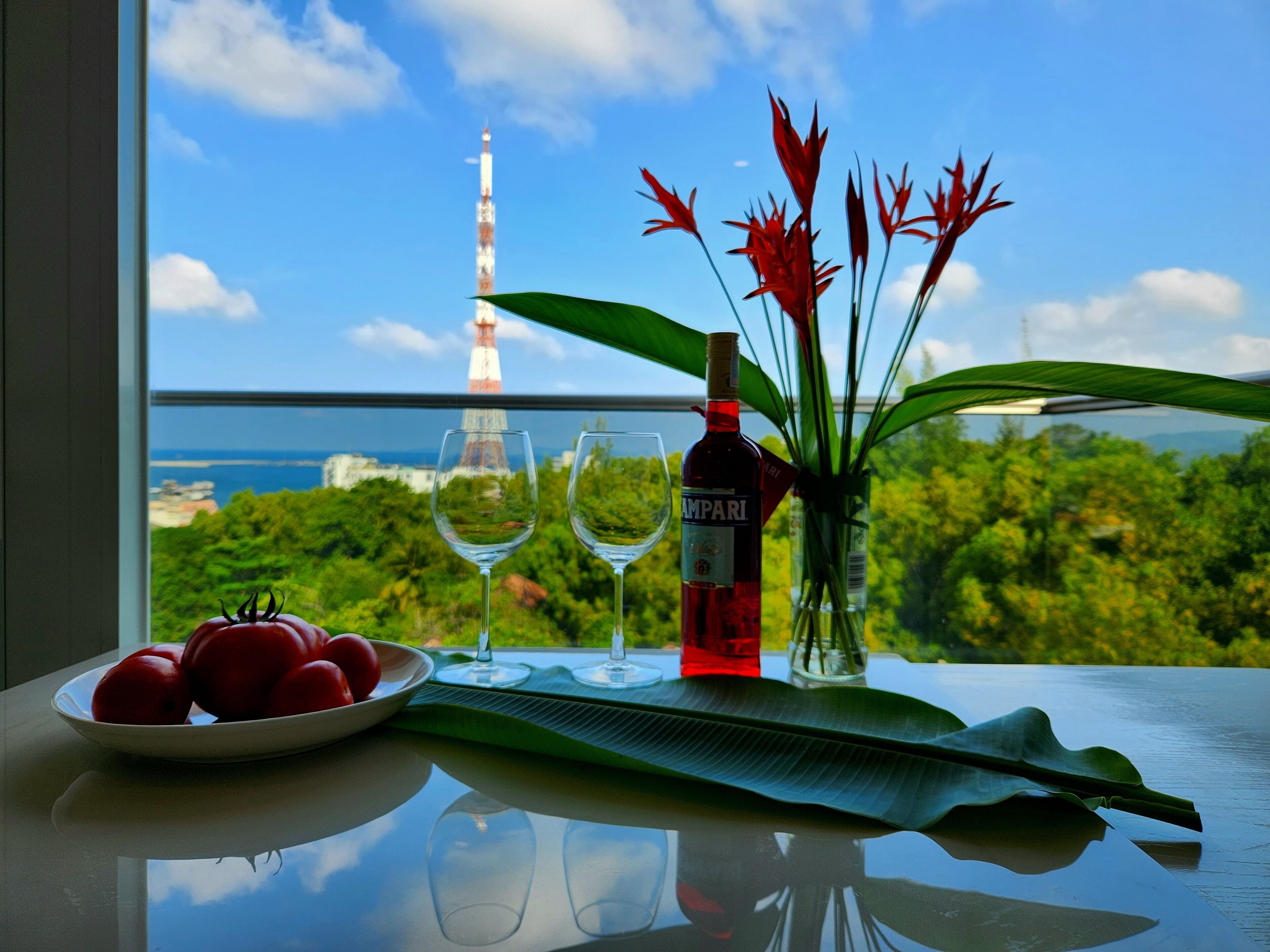 A table with a bottle of wine, two glasses, a plate of tomatoes, and a vase with red flowers, with a view of a tower and greenery.