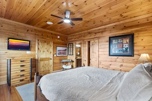 Ein Schlafzimmer in einer Holzhütte mit einem Bett, Deckenventilator, montiertem Fernseher und gerahmten Bildern an den Wänden.