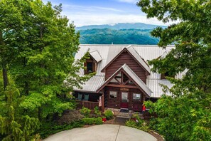 A rustic cabin with a steeply pitched roof, surrounded by lush greenery and a mountain backdrop. - Til's Den - Ask About Free Attraction Tickets! (Sevierville)