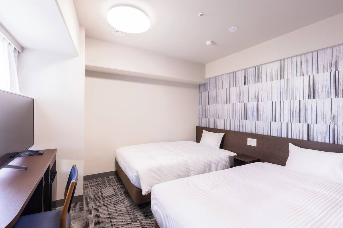 A hotel room with two beds, a desk with a chair, a television, and a patterned wall.