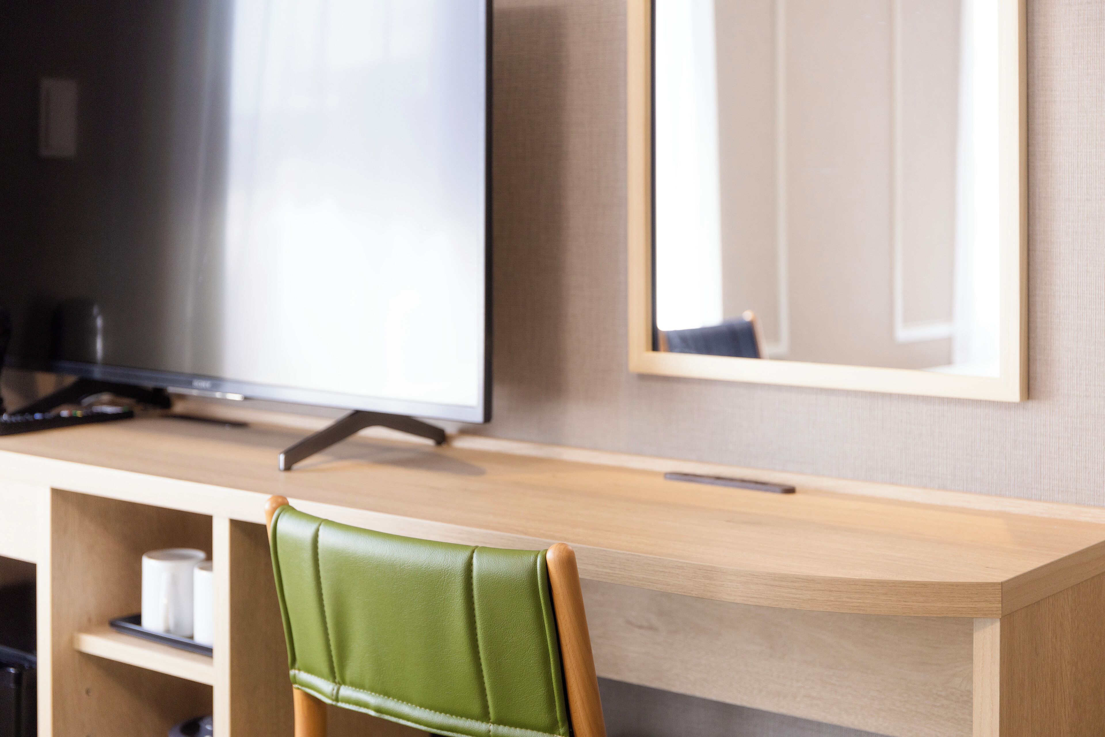 A modern hotel room with a wooden desk, a green leather chair, a flat-screen TV, and a framed mirror on the wall.