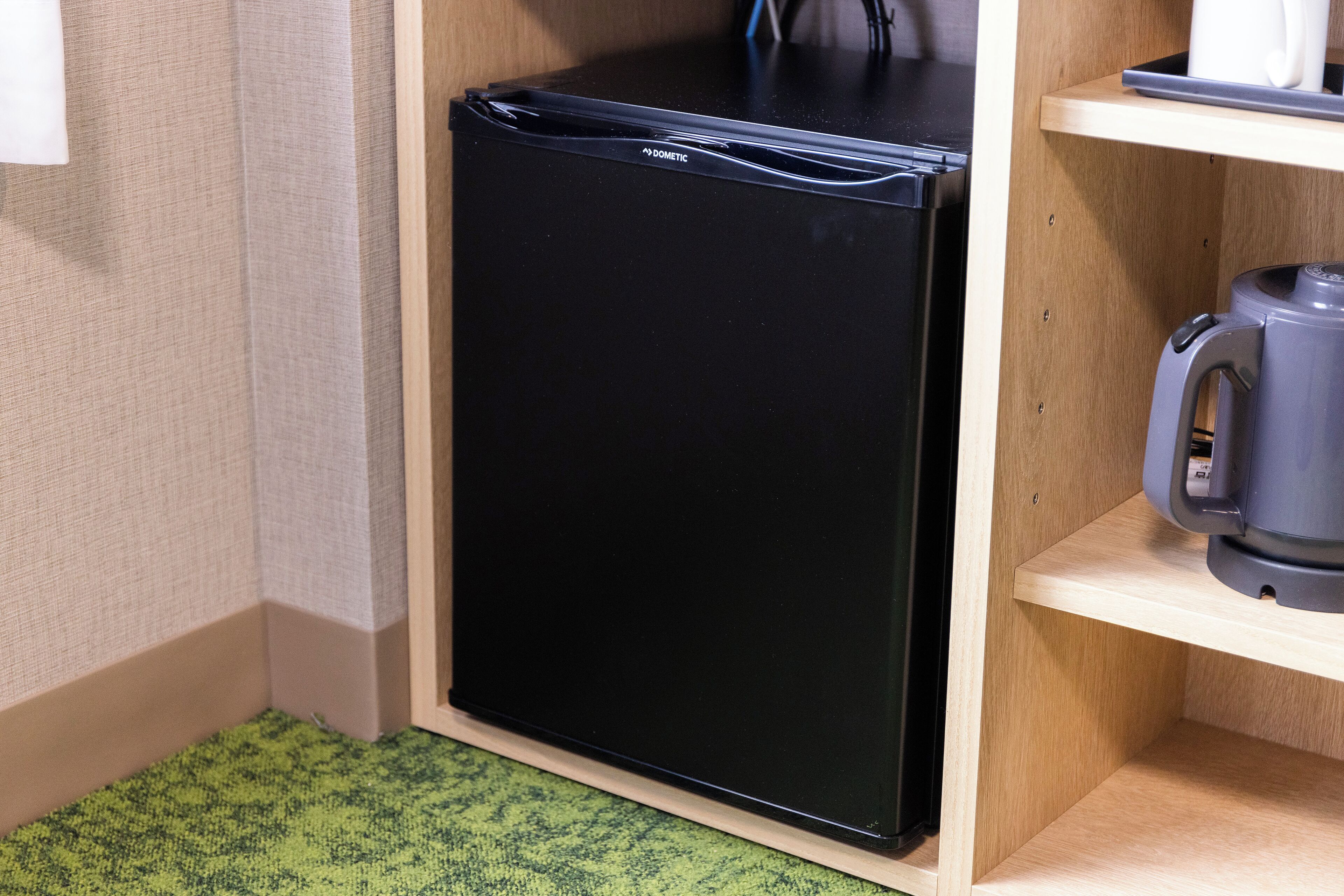Un refrigerador negro compacto en una cocina, con un estante de madera arriba y una cafetera a la derecha.