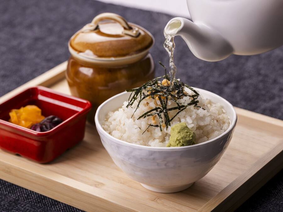 A bowl of rice topped with seaweed and green paste, a red container with yellow and purple items, and a teapot pouring liquid.