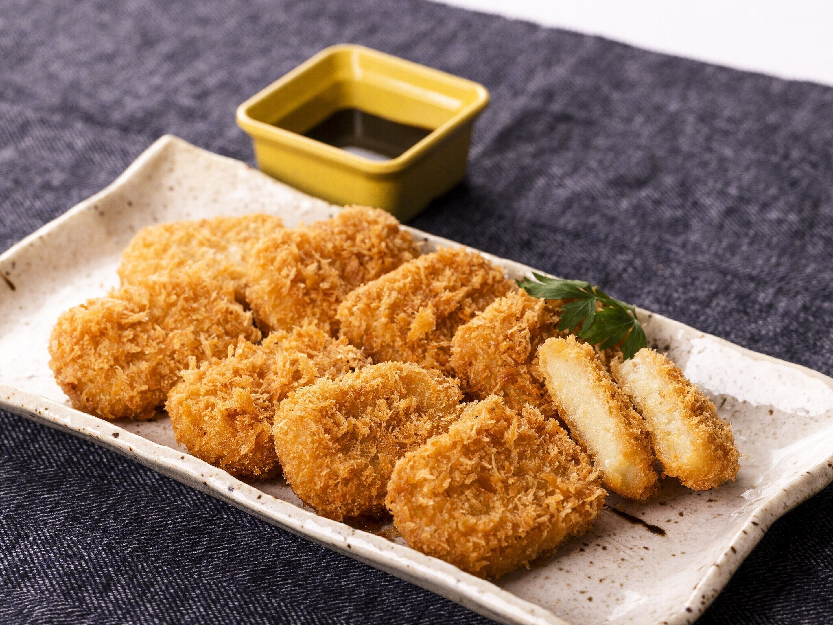 A plate of breaded and fried food items, possibly chicken, served on a rectangular ceramic plate with a small yellow dish of sauce in the background.