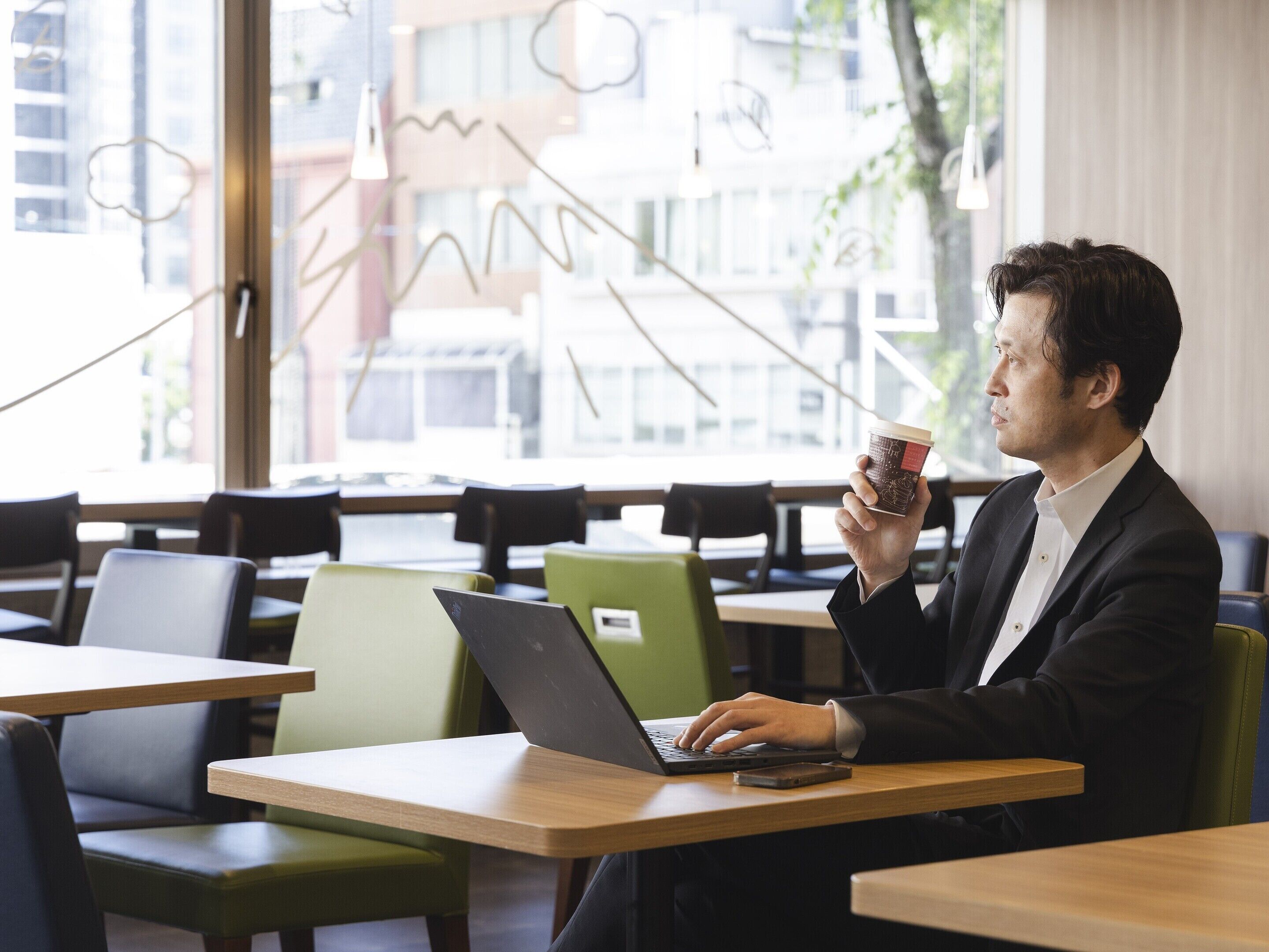 A person sitting at a table with a laptop and a coffee cup, in a cafe with large windows and modern furniture.