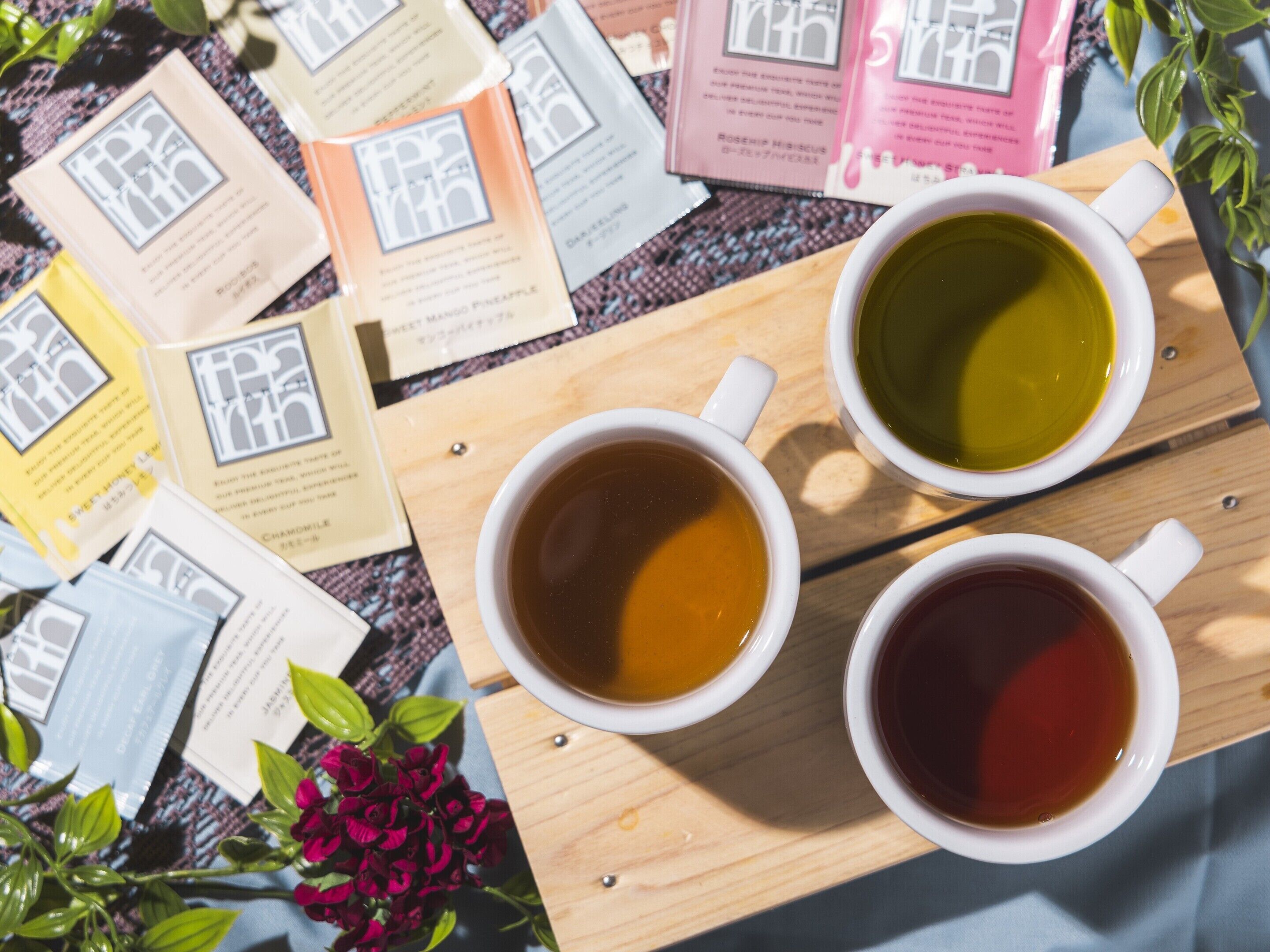 Une table en bois sur laquelle sont posées trois tasses de thé blanches, entourées de différents sachets de thé et d’un petit bouquet de fleurs.