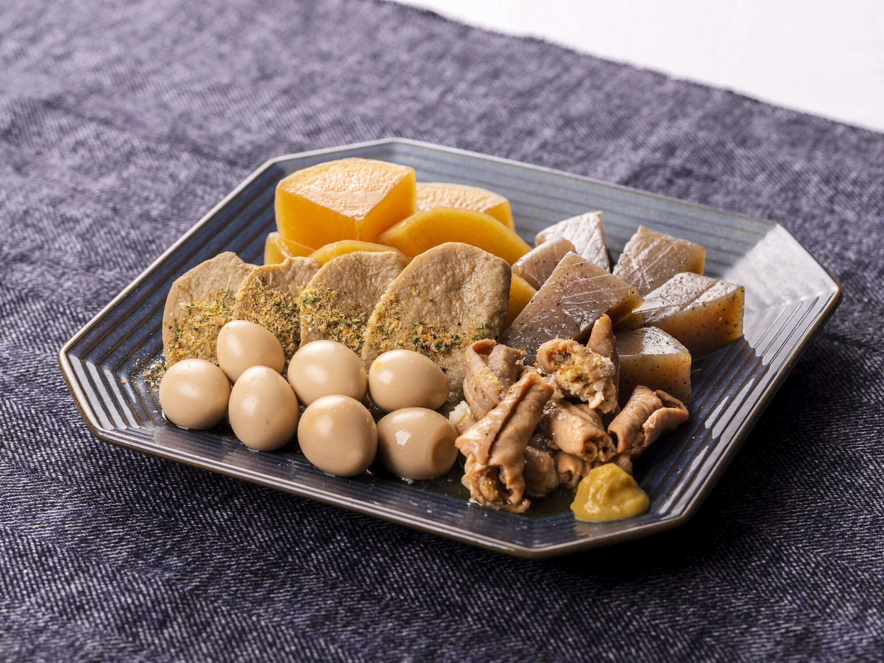 A plate of assorted food items including tofu, pickled vegetables, and possibly some meat, served on a dark textured tablecloth.