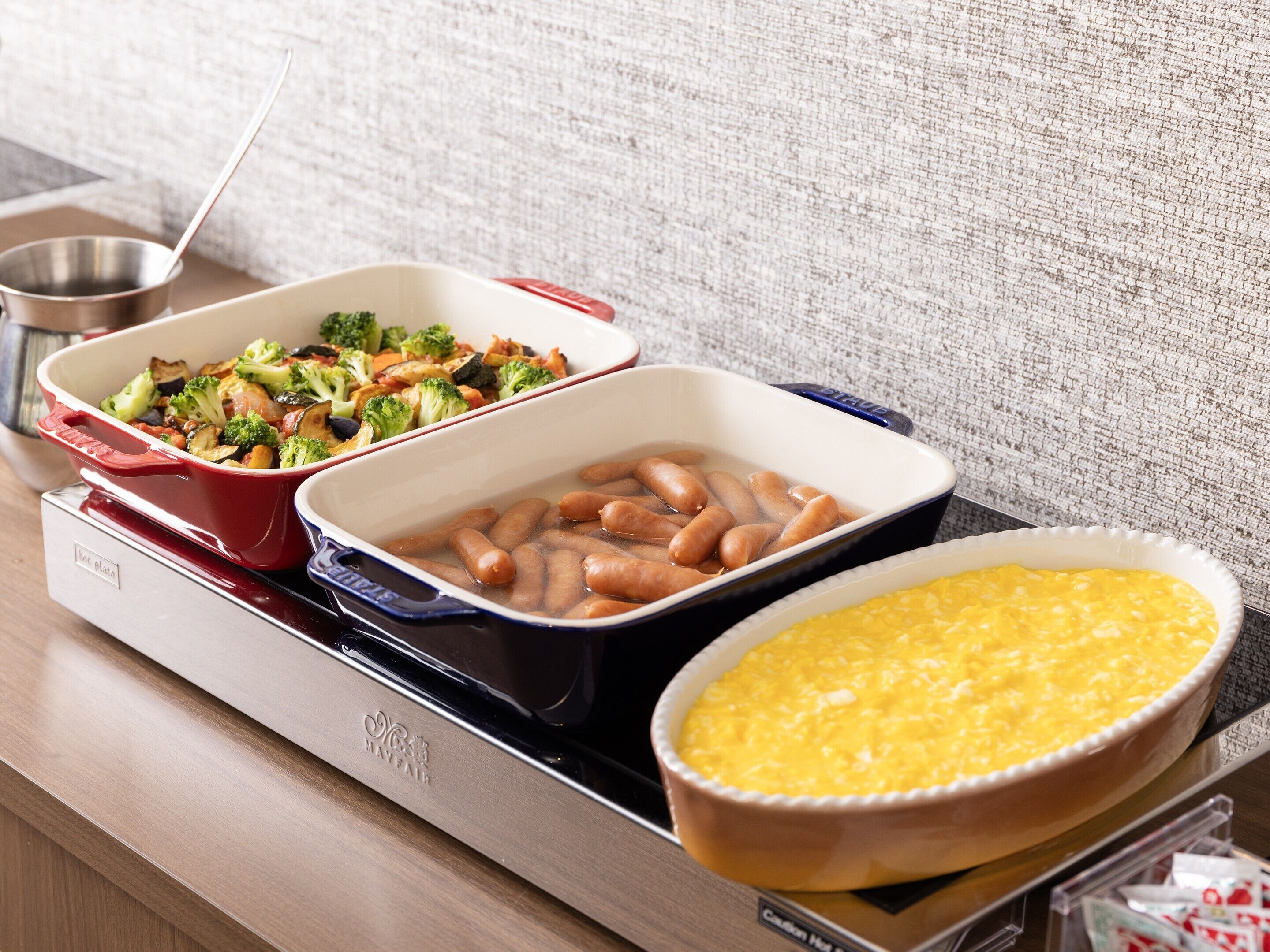 A tray with three dishes: a red casserole with vegetables, a white dish with sausages, and a yellow casserole.