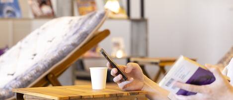A person sitting on a wooden chair with a white cushion, holding a smartphone and a book. There is a white cup on the chair.