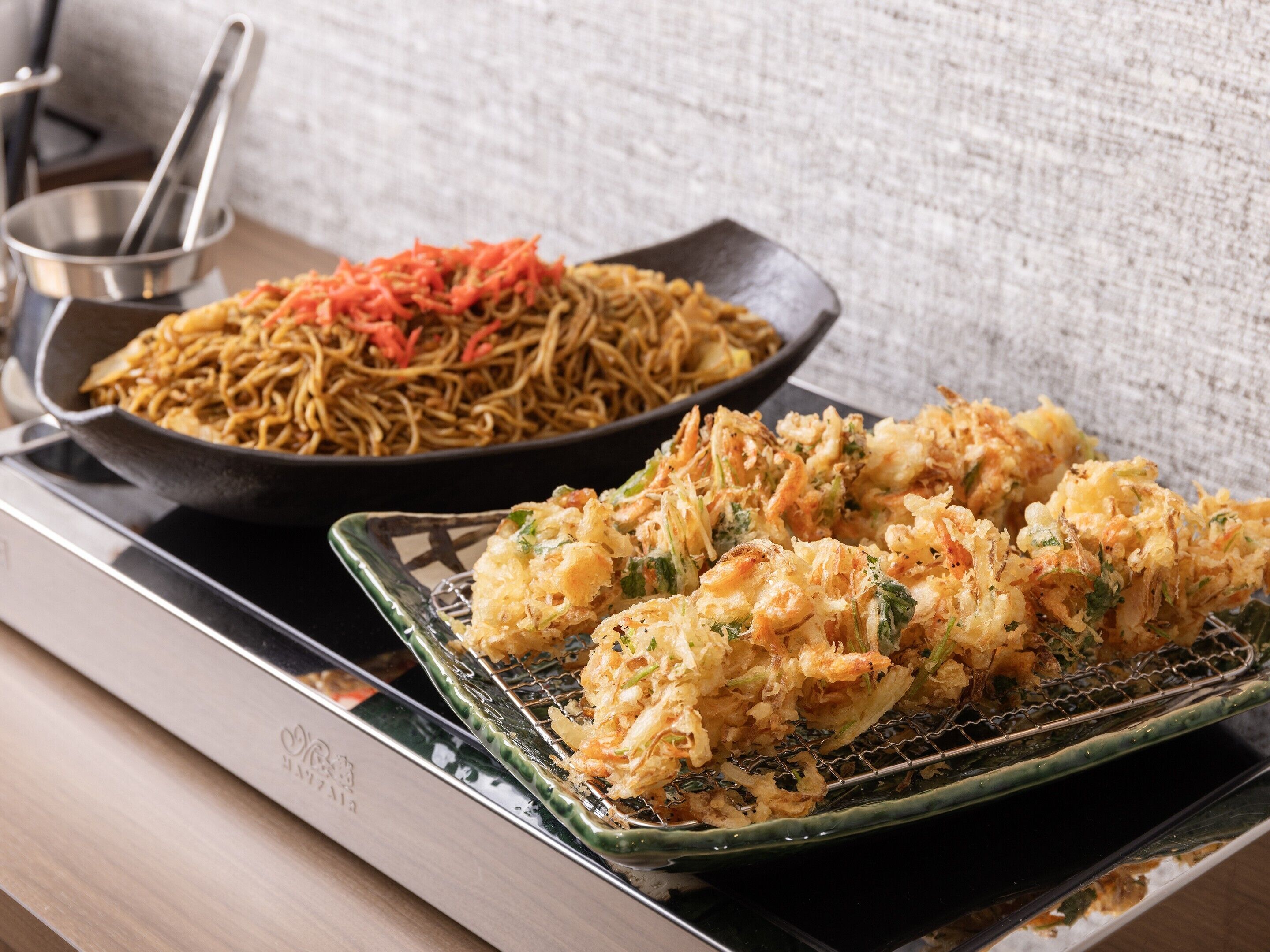 A kitchen counter with a pan of stir-fried noodles and a tray of tempura shrimp.
