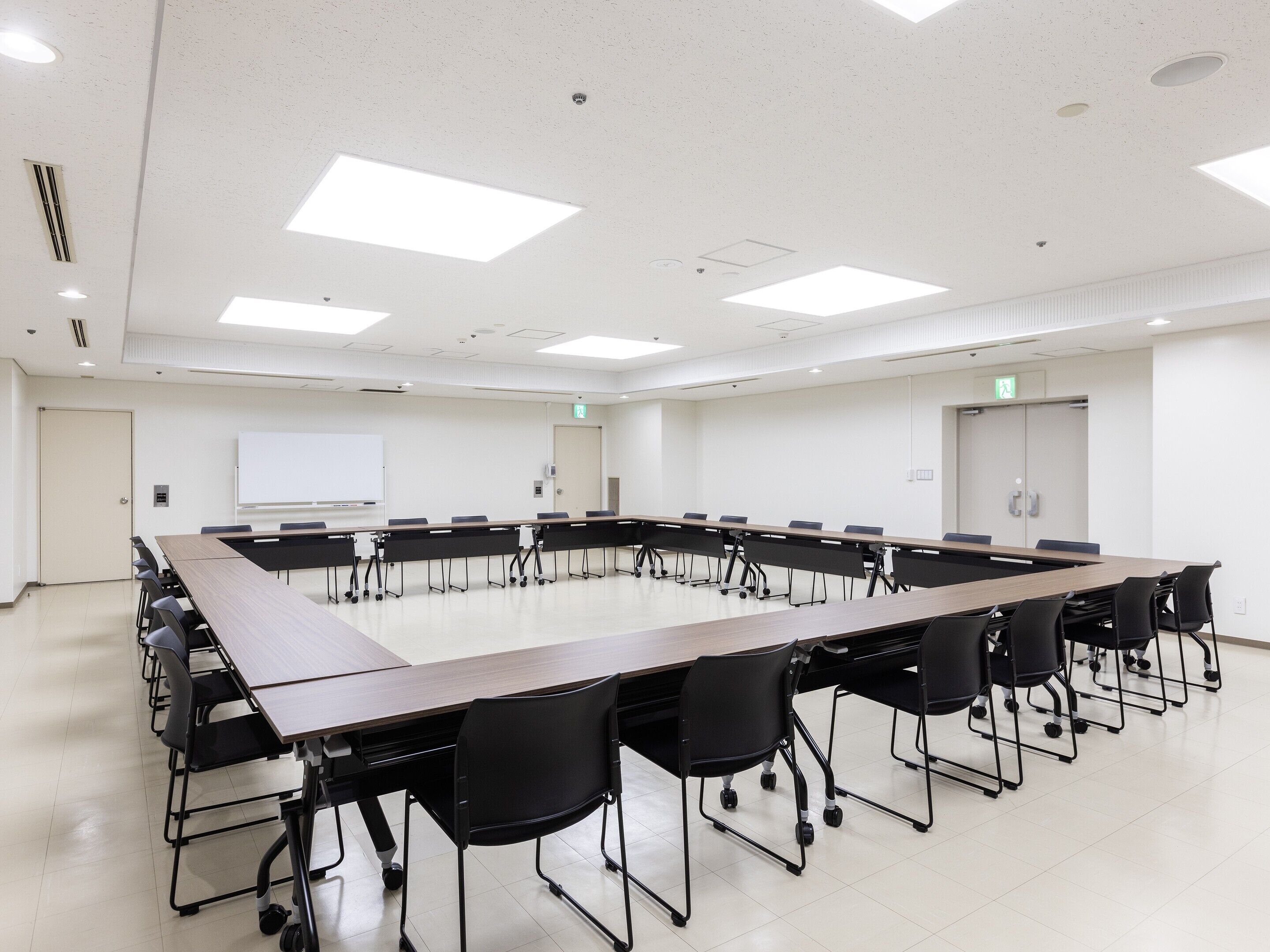 A conference room with a long table, black chairs, white walls, a whiteboard, and a door with an exit sign.