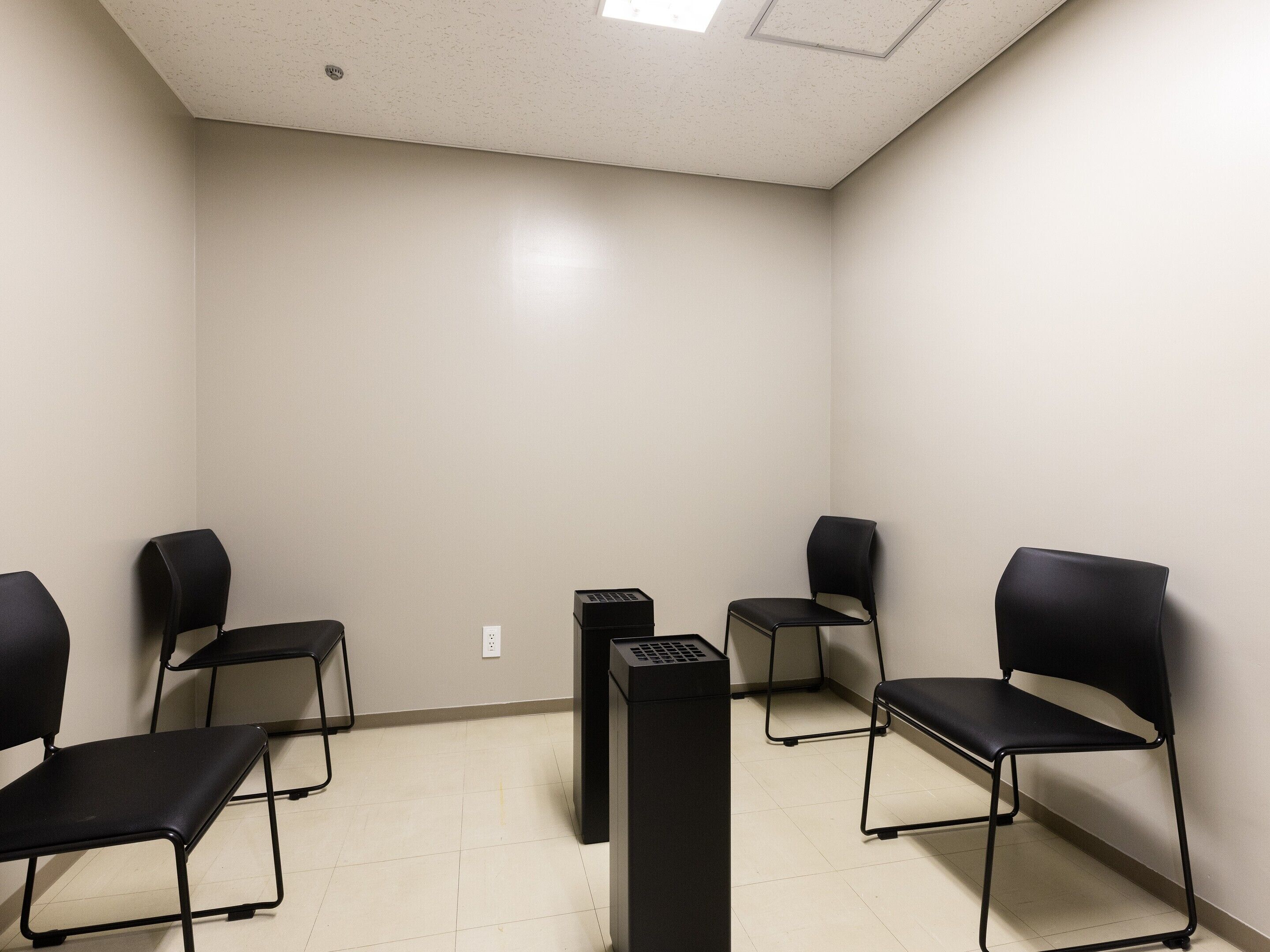 A waiting area with four black chairs arranged in a semi-circle, a black filing cabinet, and a white wall with a ceiling light.