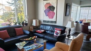 Un salón con un sofá de cuero negro, una mesa de centro de cristal con revistas, un sillón rojo y un ventanal con vistas a árboles y una casa.