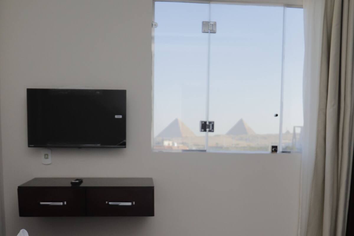 A hotel room with a bed, a TV mounted on the wall, a window with a view of pyramids, and a wooden floor.