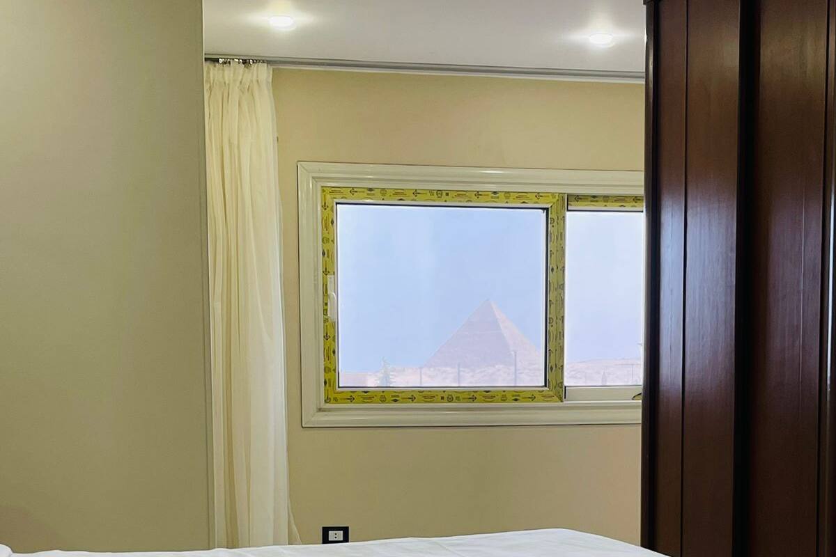 A hotel room with a bed, a window with a view of a pyramid, and a wooden wardrobe.