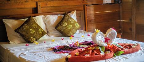Una cama bien hecha con una toalla decorativa y flores, cabecera de madera y almohadas estampadas.
