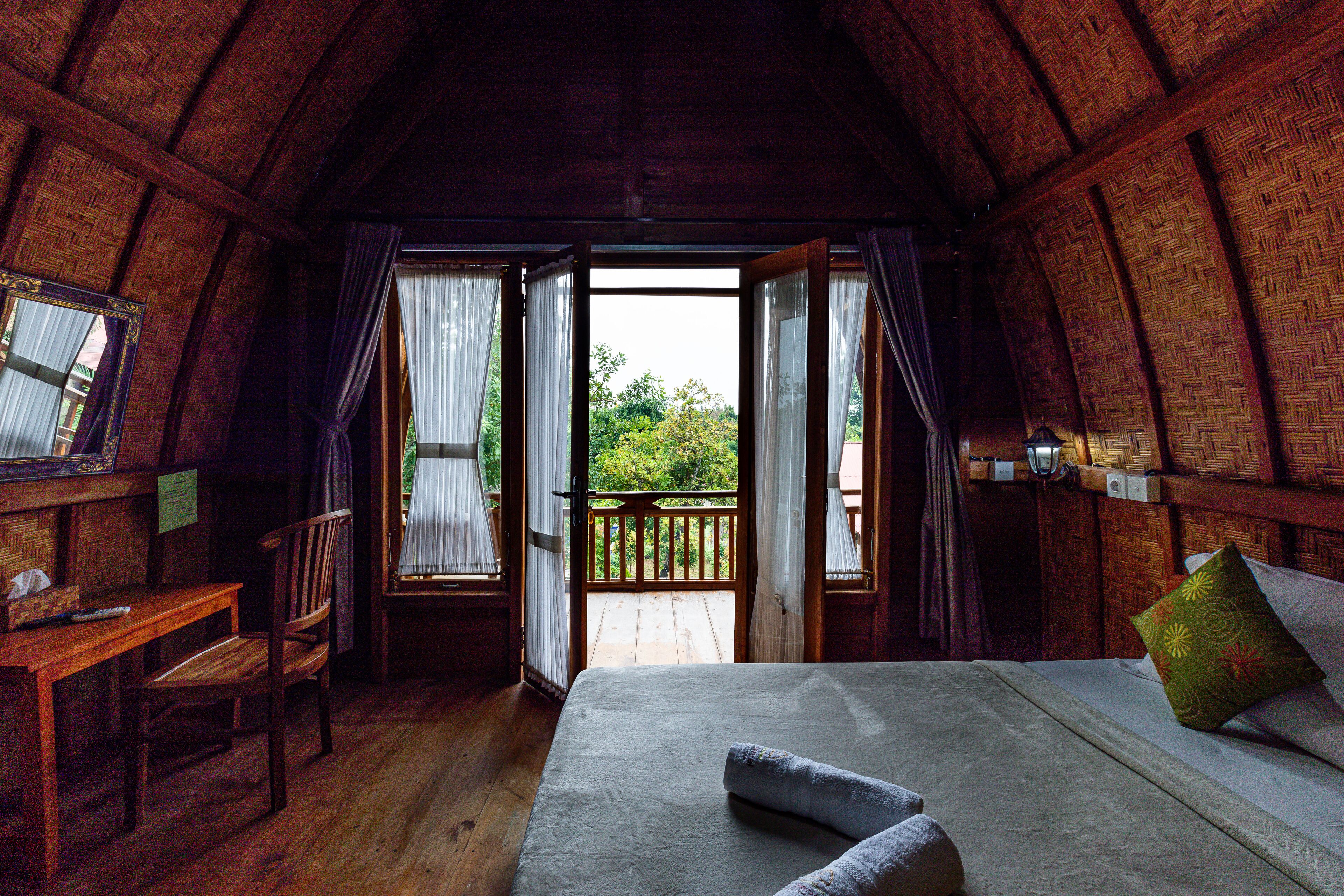 Une pièce chaleureuse avec un plafond en bois, un lit avec des oreillers, un bureau avec une chaise et une vue sur l’extérieur par une porte ouverte.
