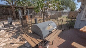 Una parrilla para barbacoa con estructura metálica y ruedas, ubicada en un patio de ladrillo con mesas de picnic de madera.