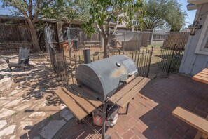 Un barbecue avec une structure en métal et des roulettes, installé sur une terrasse en briques avec des tables de pique-nique en bois.