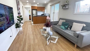Una sala de estar moderna con un sofá gris, una mesa de centro y un televisor. Se ve una cocina al fondo.