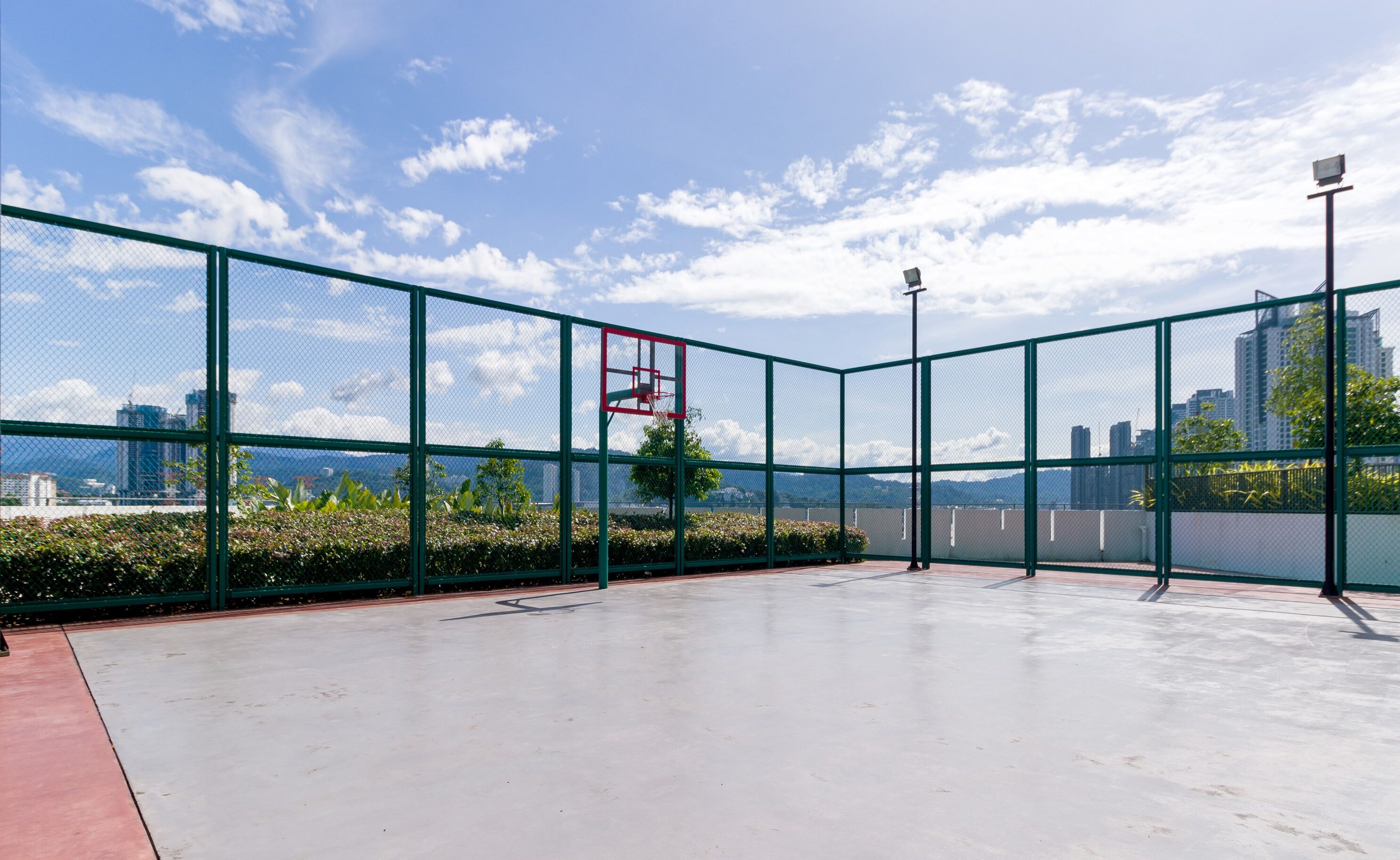 An outdoor basketball court with a net, surrounded by a green fence, and a cityscape in the background.