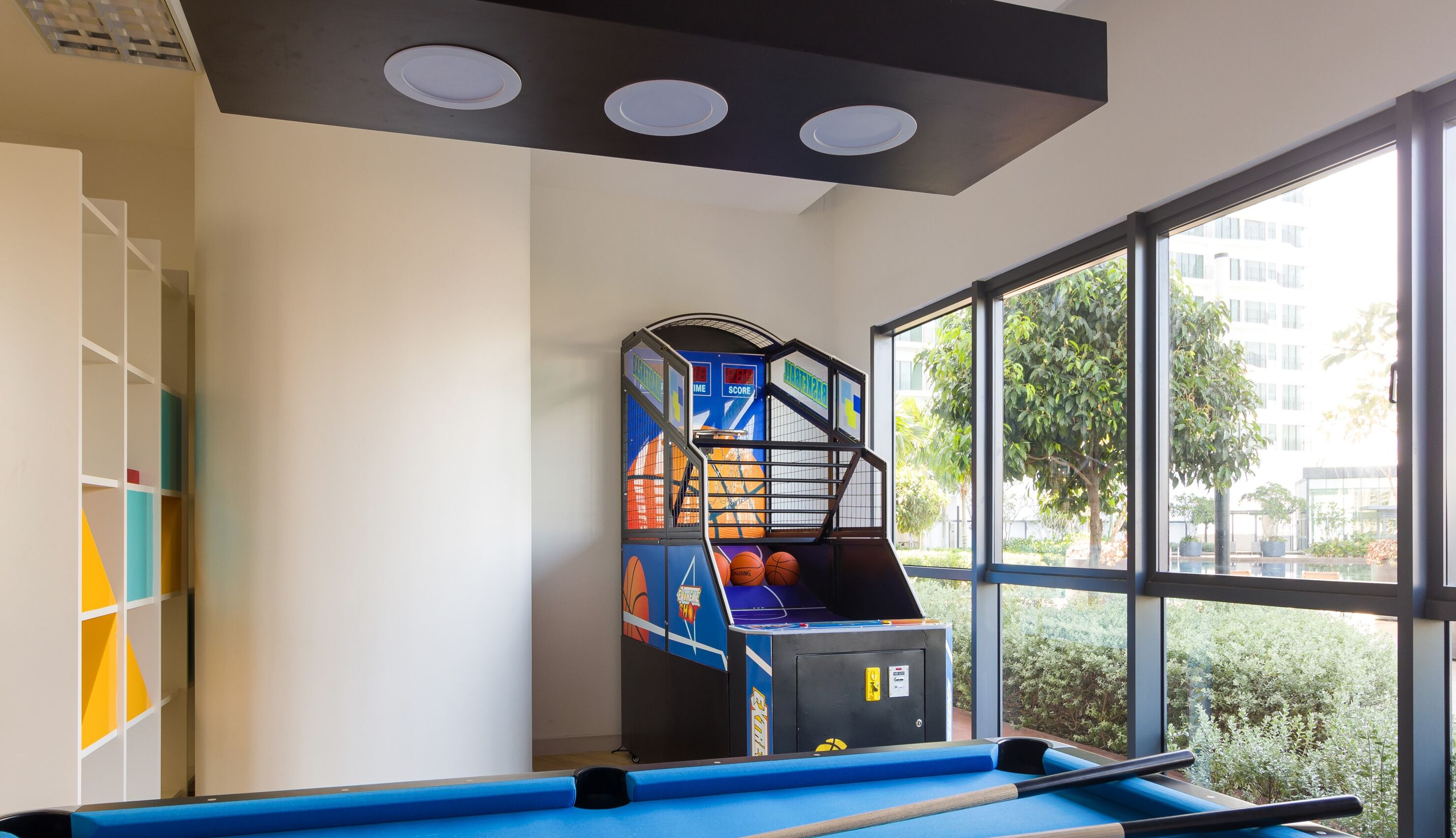 A pool table with a basketball arcade game, a window with a view of greenery, and a white wall with a geometric pattern.