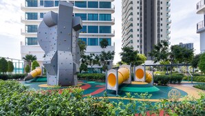 Um parquinho moderno com estruturas de escalada e escorregadores, rodeado por áreas verdes e edifícios altos.