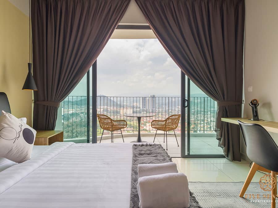 A hotel room with a bed, a desk, two chairs, a balcony with a table and chairs, and a view of the cityscape.