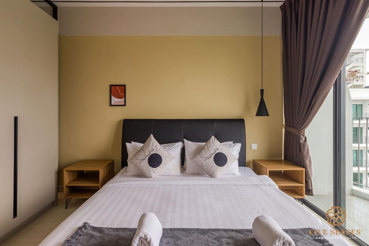 A modern hotel room with a large bed, two nightstands, a wooden side table, a single hanging light, and a view of the outside through a window with curtains.
