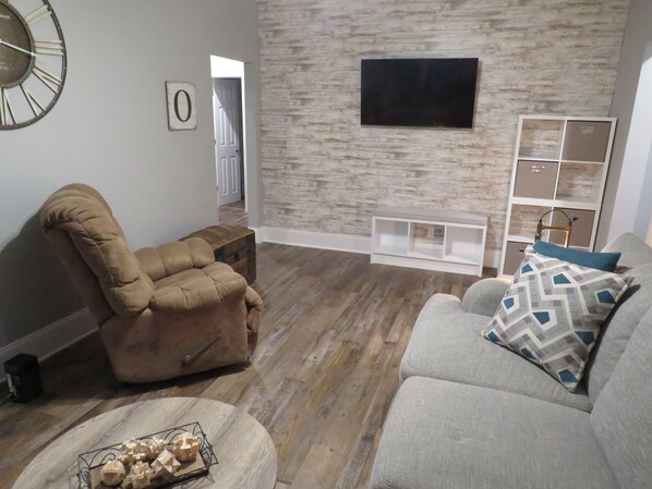 Una sala de estar con un sillón reclinable marrón, un sofá gris con almohadones estampados, una mesa de centro de madera y un televisor de pantalla plana montado en una pared con diseño de ladrillo.